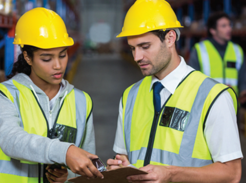 Two people wearing high vis and hard hats discussing information over a clipboard in a warehouse