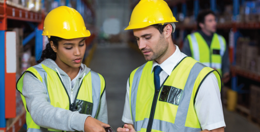 Two people wearing high vis and hard hats discussing information over a clipboard in a warehouse