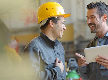 Smiling man holding a tablet talking to another smiling man wearing a hard hat in a warehouse