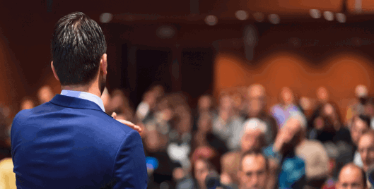 Back of a man in a business suit speaking to a crowd of people at an event