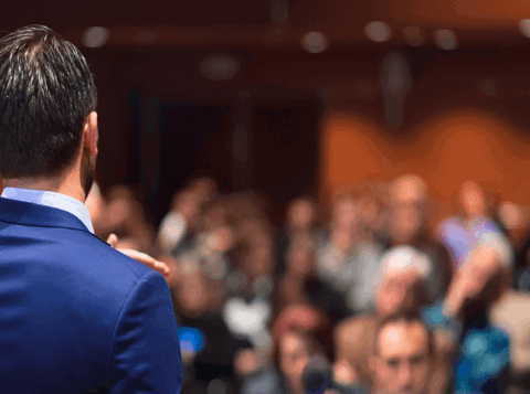 Back of a man in a business suit speaking to a crowd of people at an event