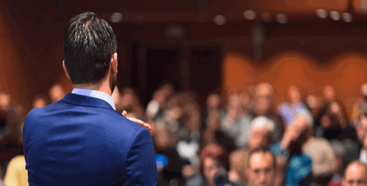 Back of a man in a business suit speaking to a crowd of people at an event
