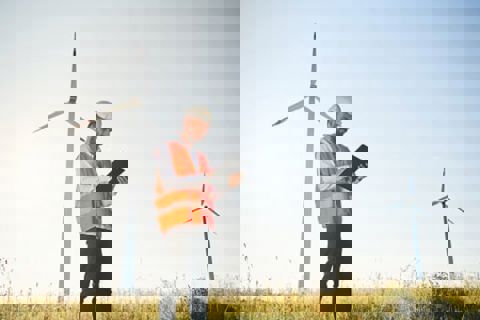 Man in high vis and hard hat holding a clipboard standing in front of a wind turbine