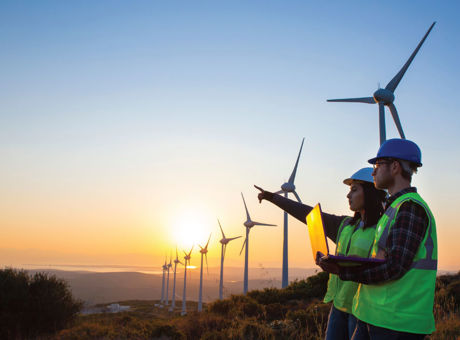 Two people wearing high vis and PPE stood in a wind turbine field holding a laptop and pointing to the distance